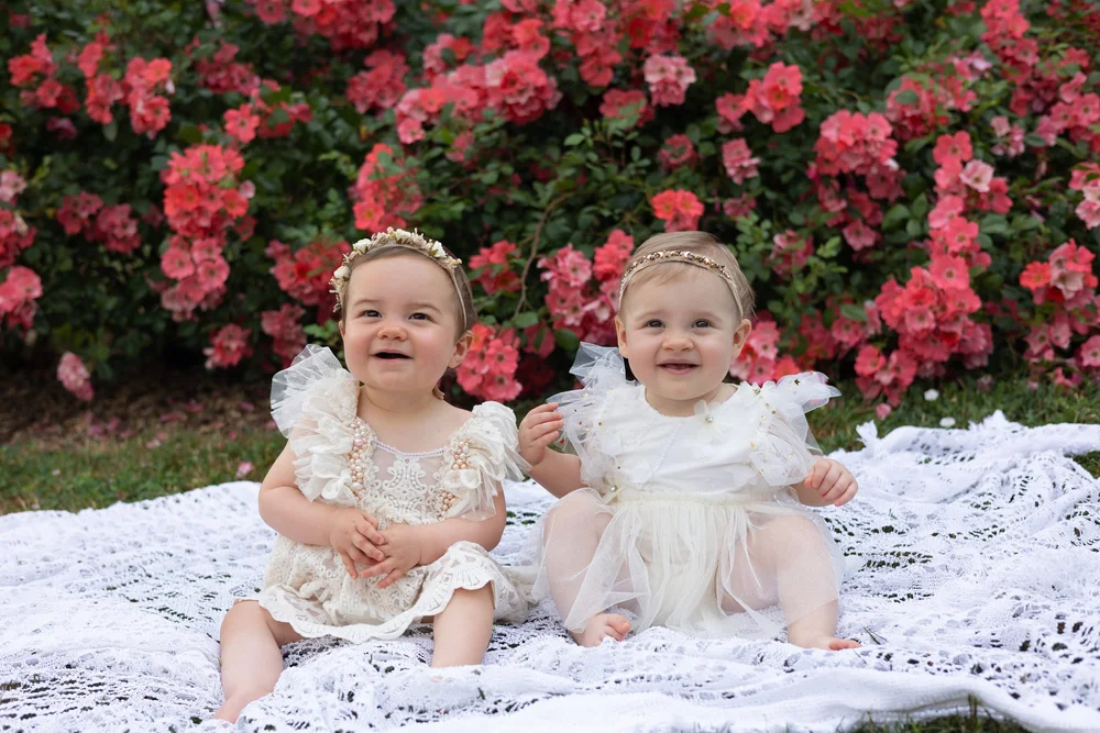 Deux bébés en robe de dentelle assis devant des roses rouges.