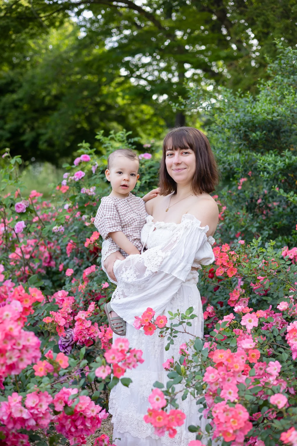 Maman en robe blanche portant son bébé au milieu de rosiers roses.