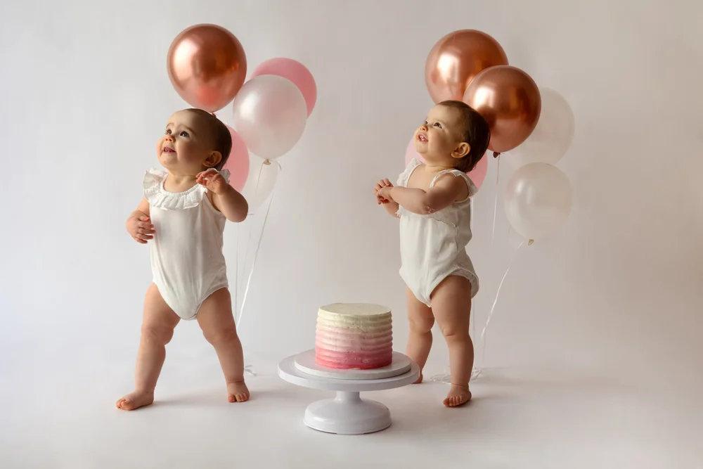 Jumeaux en body blanc avec des ballons cuivrés autour d’un gâteau.