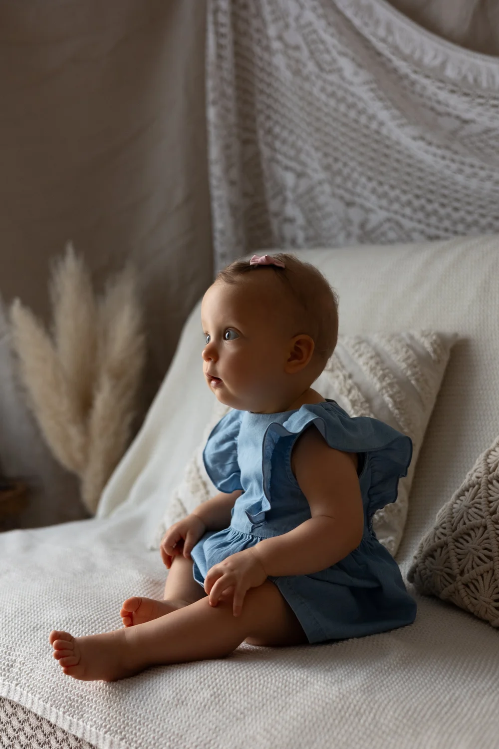 Bébé en salopette bleue assise sur un lit, devant des pampas.