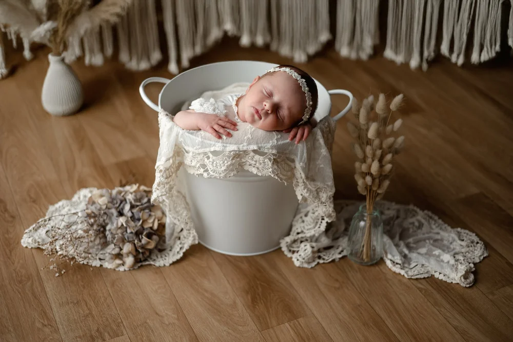 Nouveau-né endormi dans un seau en métal garni de dentelle, sur un parquet.