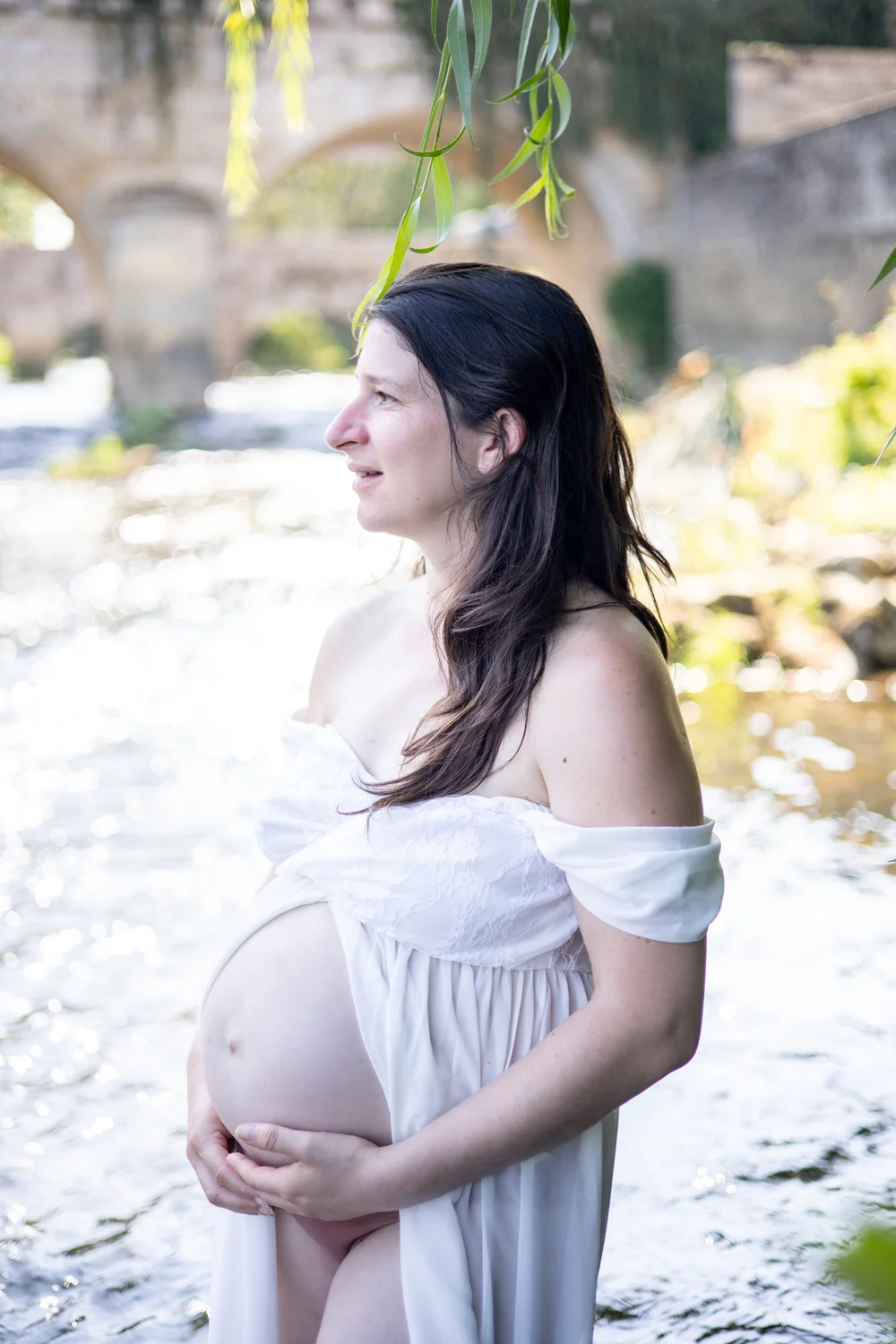 Femme enceinte en robe blanche aux épaules dénudées, au bord d’une rivière.
