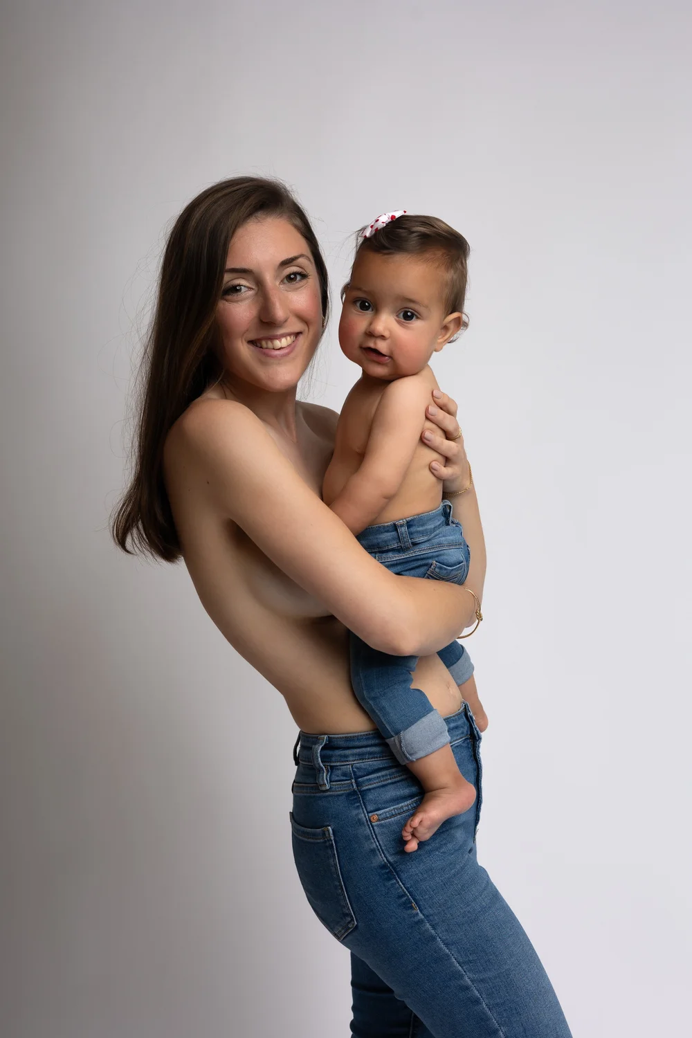 Maman en jean tenant son bébé contre elle, sur fond clair.