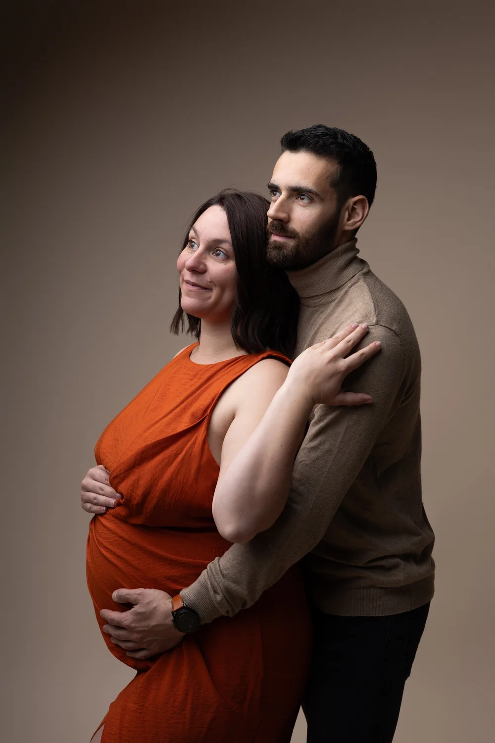 Couple en studio, la femme enceinte en robe orange, son conjoint derrière elle.