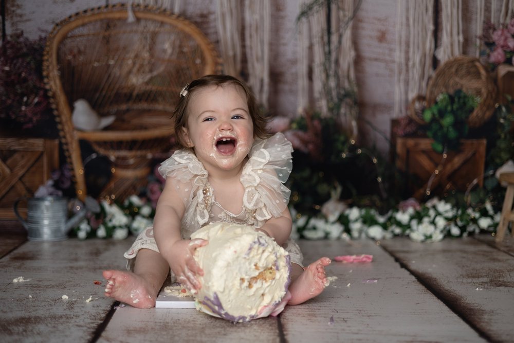 Bébé riant en robe de dentelle avec son gâteau, décor blanc.