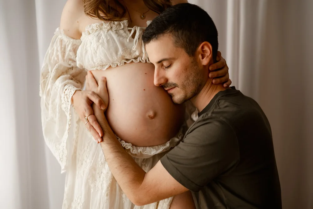 Futur papa enlaçant le ventre rond de sa compagne en robe de dentelle crème.