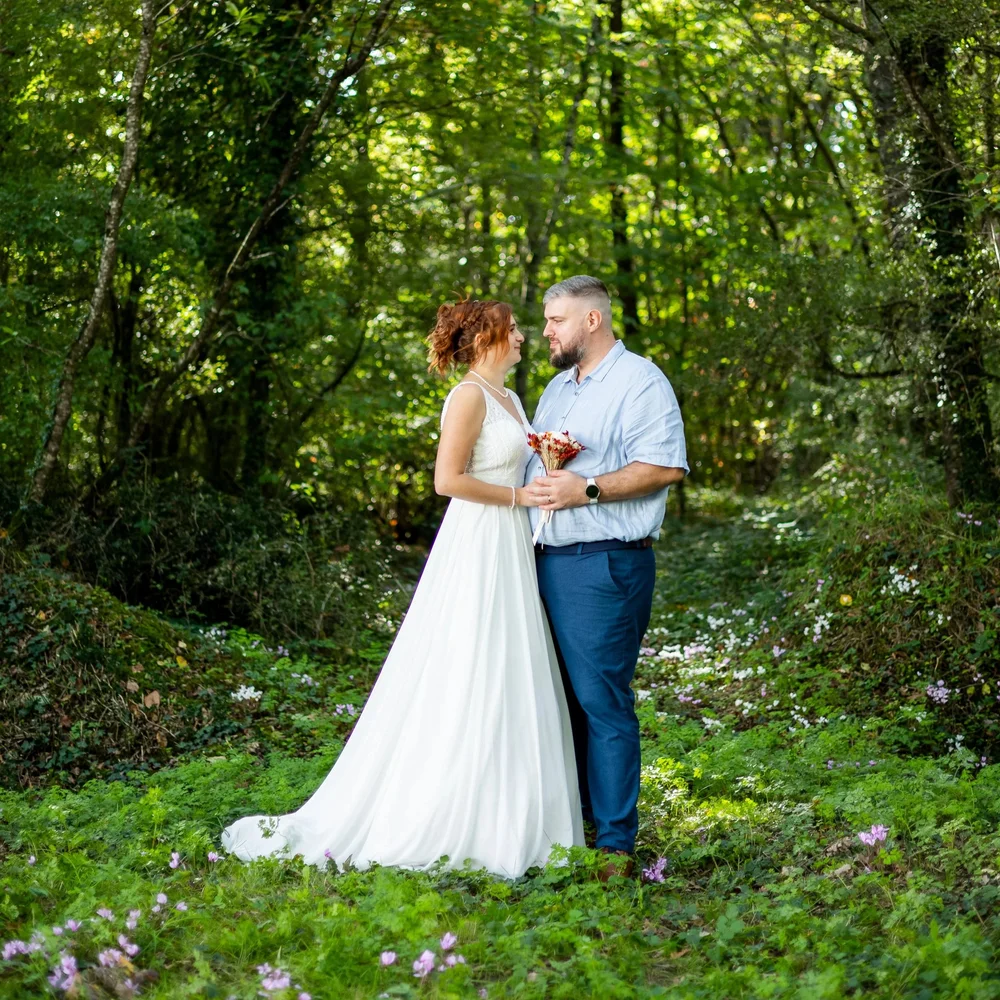 Couple de mariés face à face dans un sous-bois, la mariée tenant un petit bouquet.