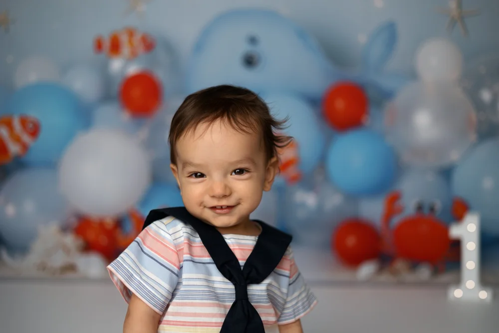 Bébé en tenue de marin devant un décor de ballons bleus et de poissons.