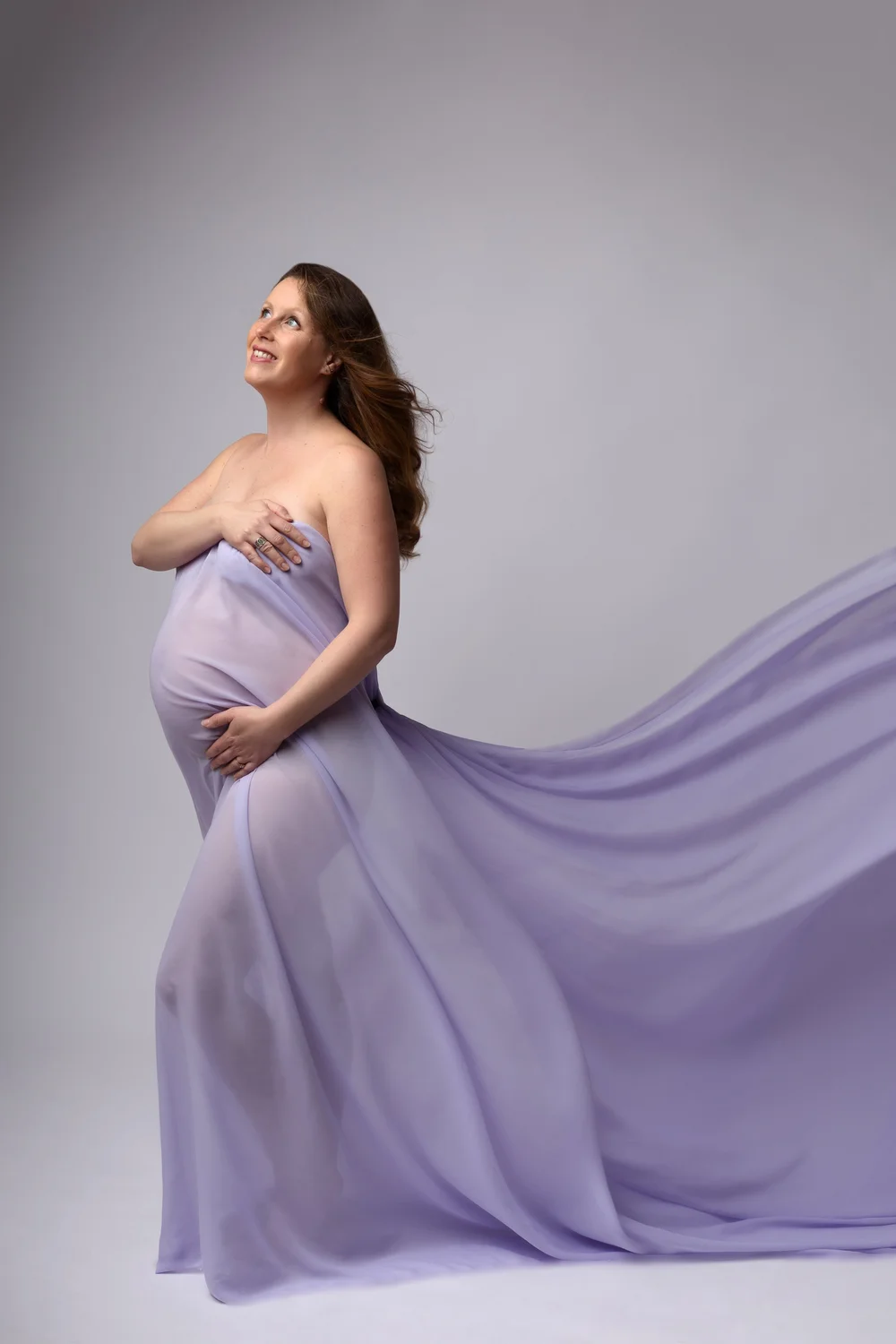 Femme enceinte drapée d’un voile lilas, de profil, sur fond clair.