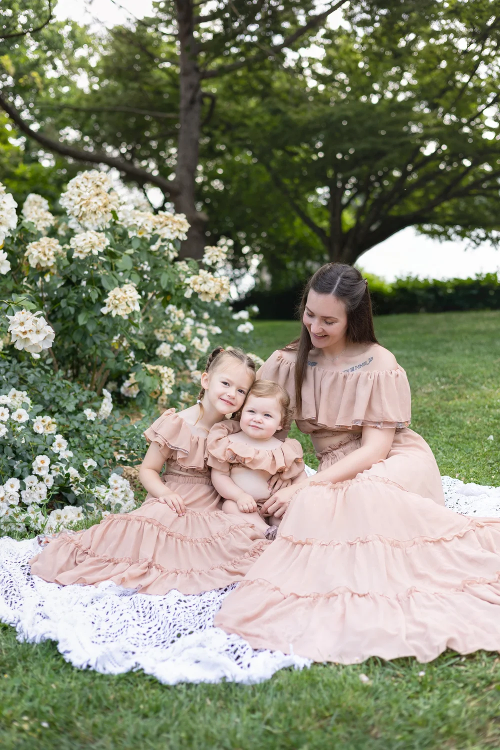 Maman assise sur un plaid avec ses deux enfants, sous de grands arbres.