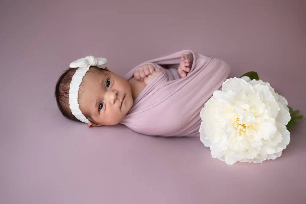 Nouveau-né emmailloté de rose avec un bandeau, près d’une pivoine.