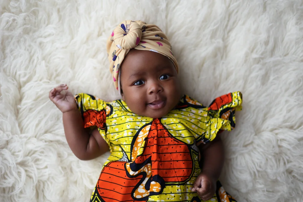 Bébé en robe wax colorée et turban, allongée sur une fourrure.