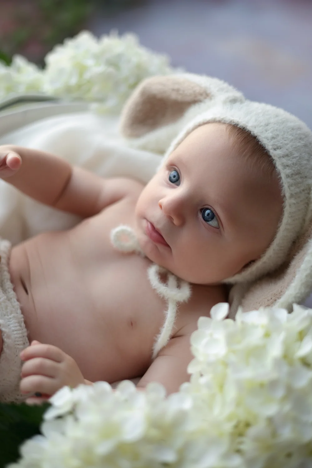 Bébé en bonnet de lapin allongé près d’hortensias blancs.