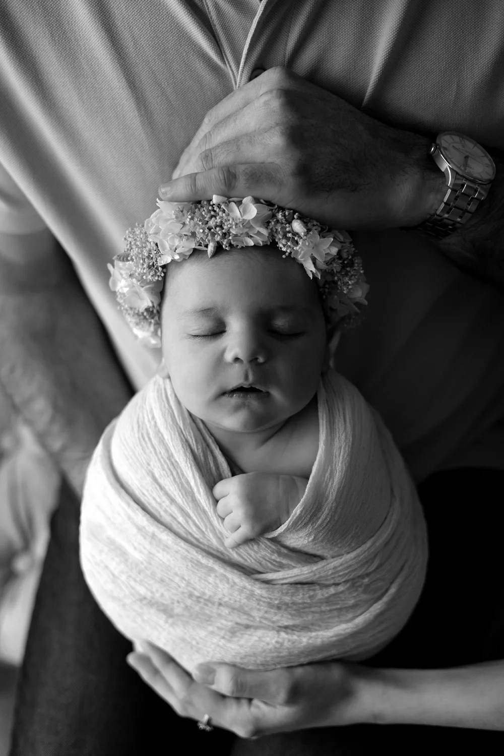 Photo en noir et blanc : nouveau-né emmailloté, couronne de fleurs, dans les mains de ses parents.