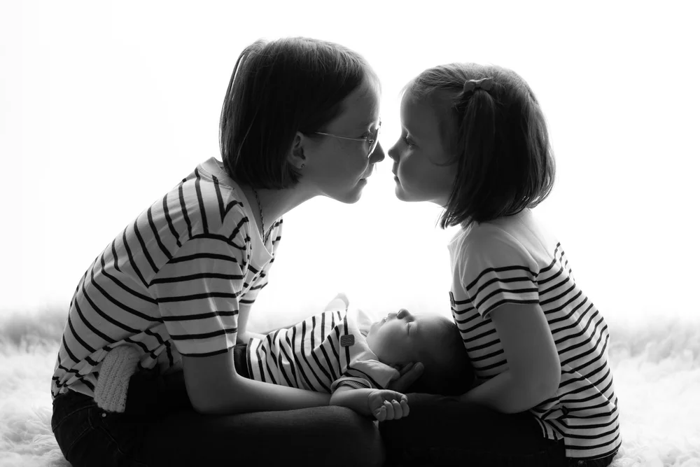 Maman et grande sœur nez à nez au-dessus du nouveau-né, en noir et blanc.