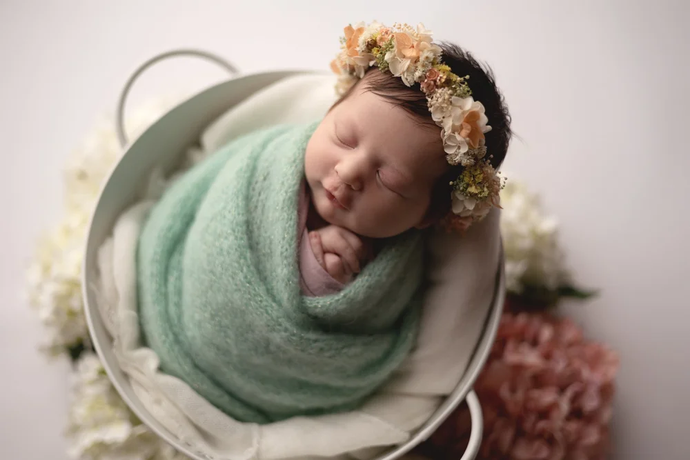 Nouveau-né emmailloté de vert d’eau, couronne de fleurs, dans un seau.