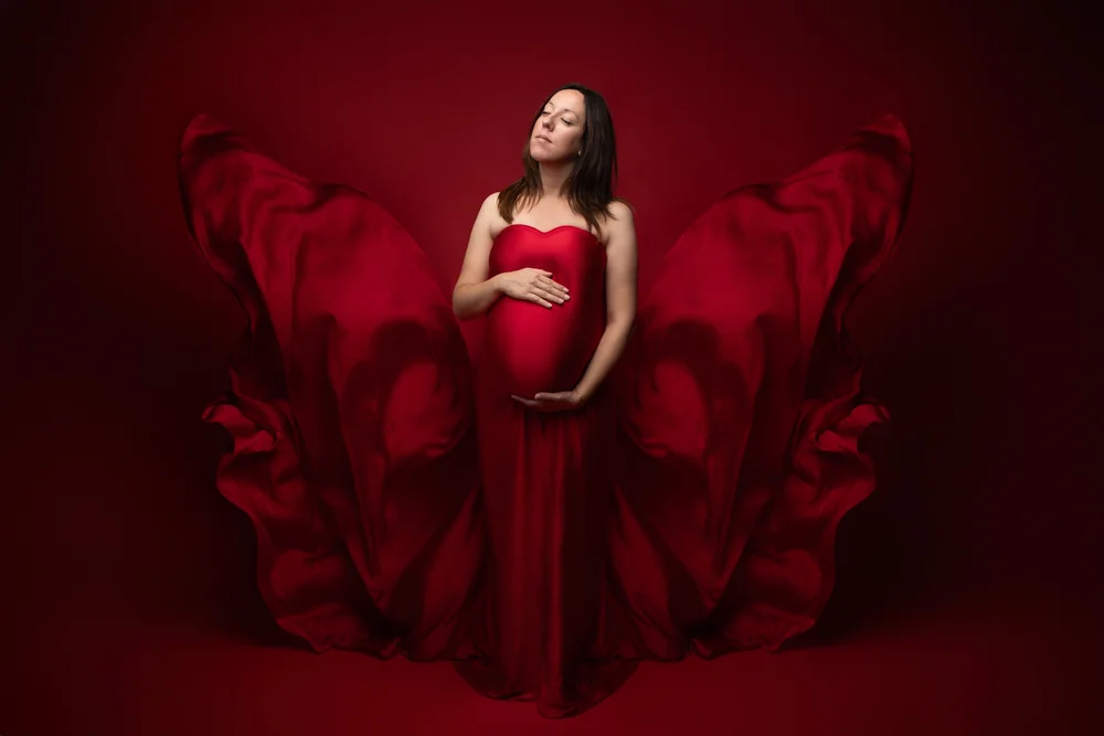 Femme enceinte en robe rouge dont le voile vole, sur fond rouge.
