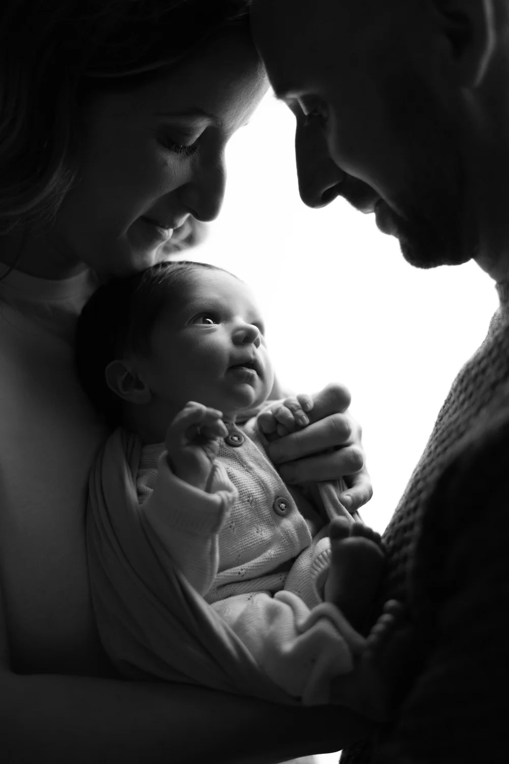 Parents penchés sur leur nouveau-né, en noir et blanc, en contre-jour.