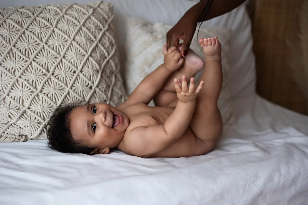 Bébé riant allongé sur un lit, sa maman lui tenant les pieds.