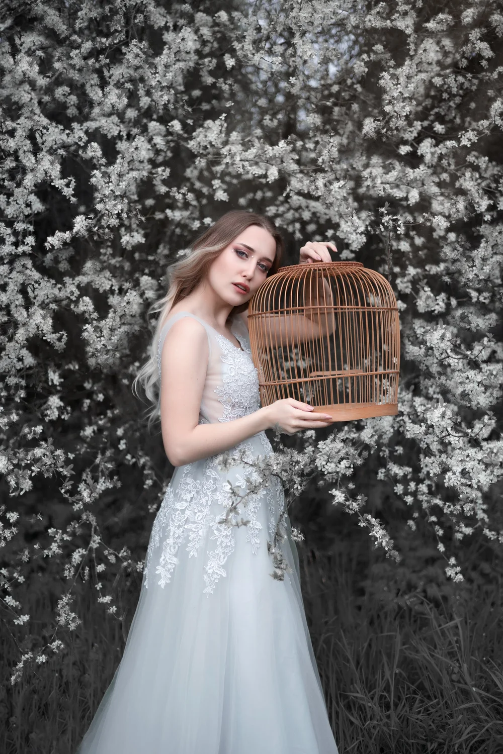 Femme en robe blanche tenant une cage à oiseaux, parmi des fleurs blanches.