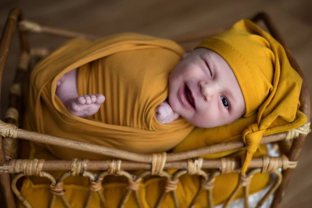 Nouveau-né souriant en bonnet moutarde dans un petit lit en rotin.