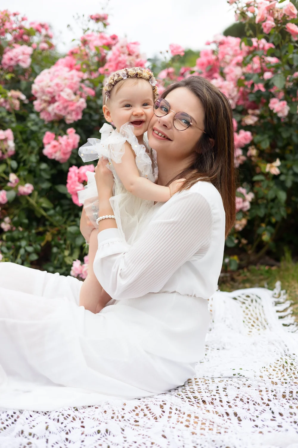Maman en robe blanche tenant son bébé devant des roses.