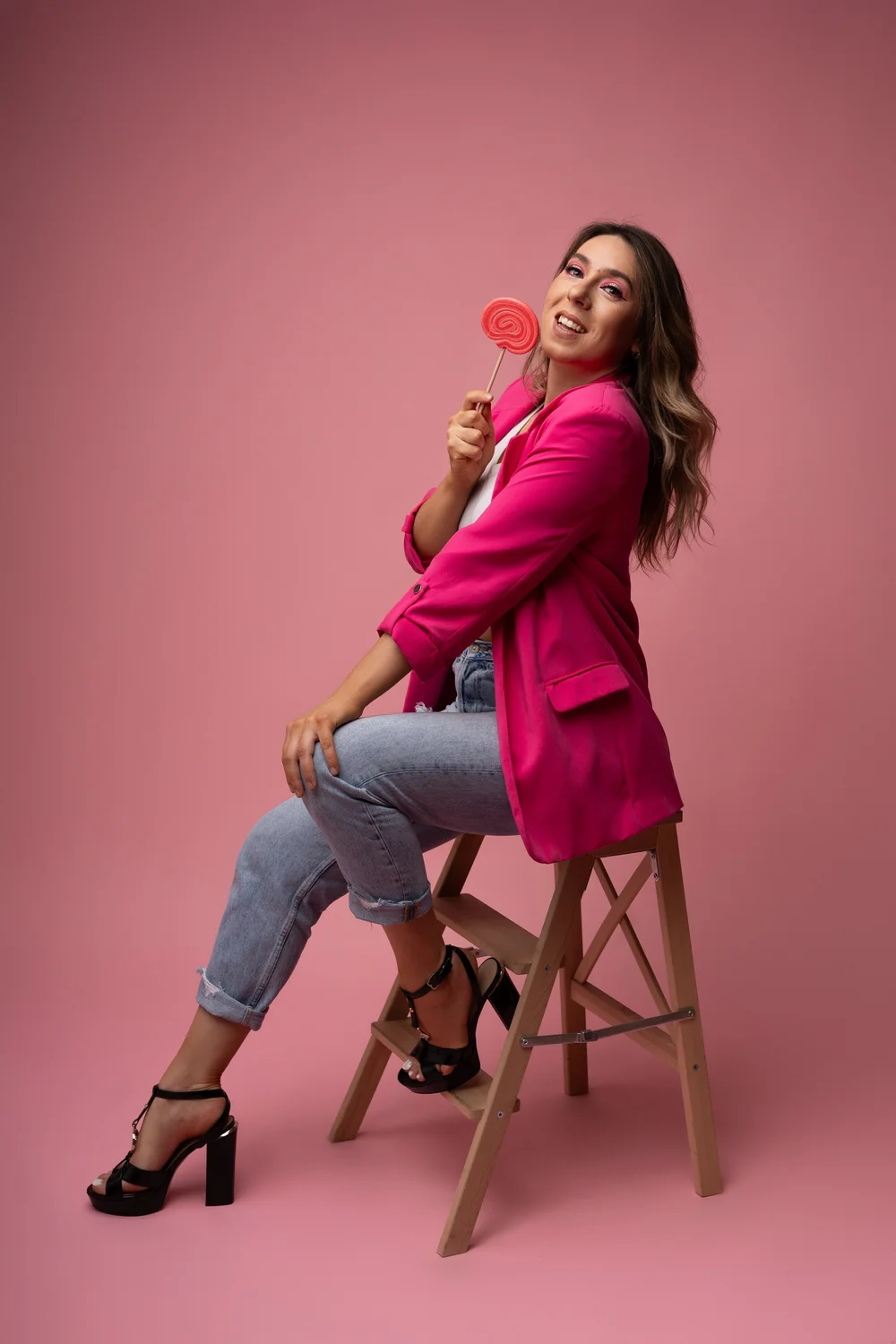 Femme en veste rose tenant une sucette, assise sur un tabouret, fond rose.