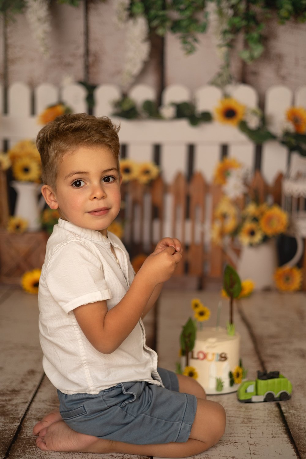 Petit garçon assis devant un décor de tournesols et de barrière blanche.