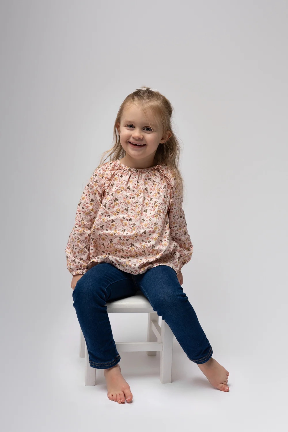 Petite fille en blouse fleurie assise sur un tabouret, en studio.