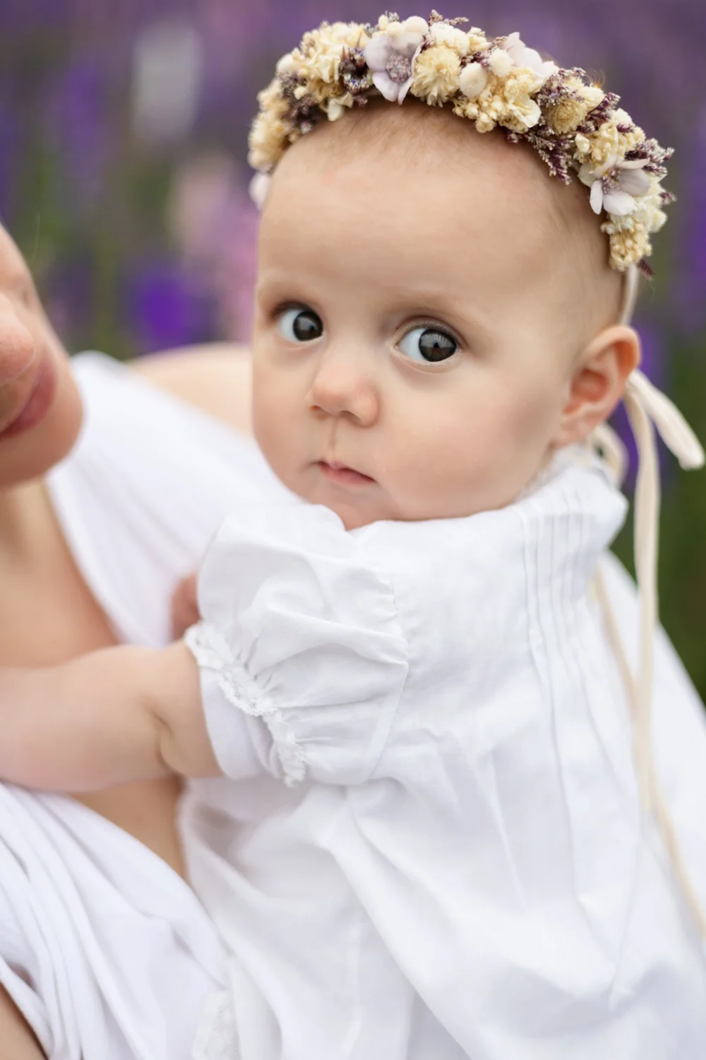 Bébé avec une couronne de fleurs dans les bras de sa maman, parmi des fleurs violettes.
