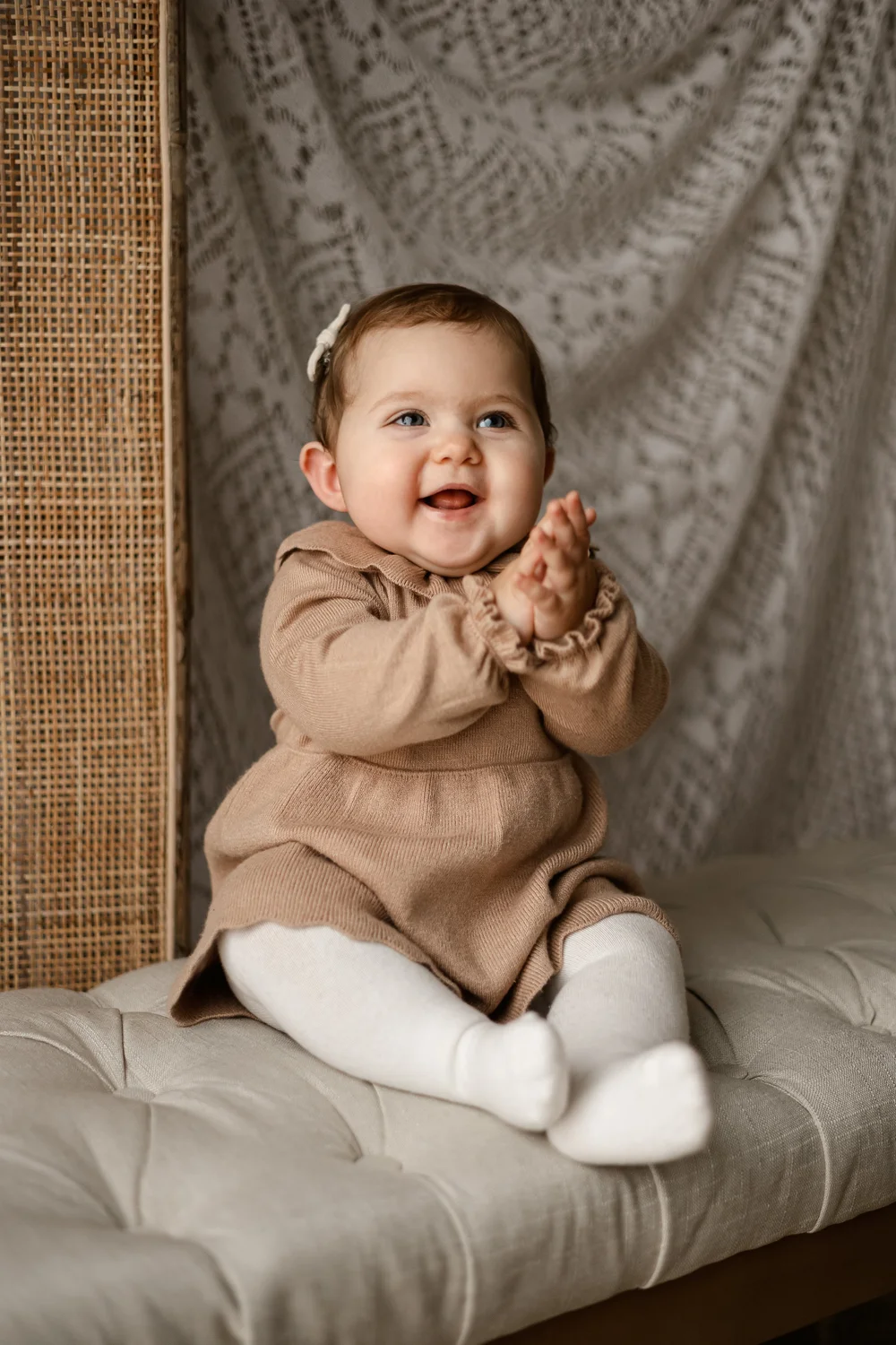 Bébé souriante en robe beige assise devant un paravent en rotin.
