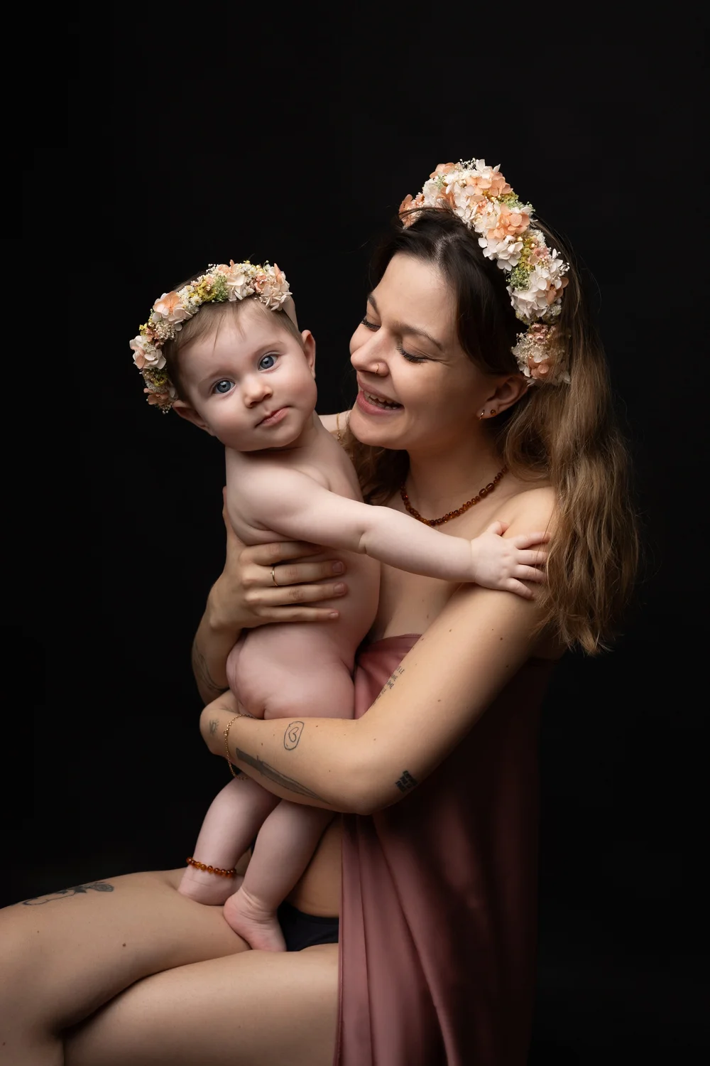 Maman et bébé couronnés de fleurs, joue contre joue, sur fond noir.