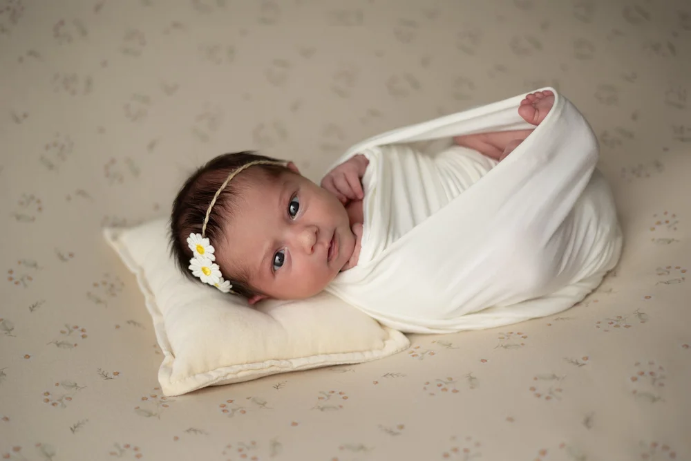 Nouveau-né emmailloté de blanc les yeux ouverts, sur un coussin.