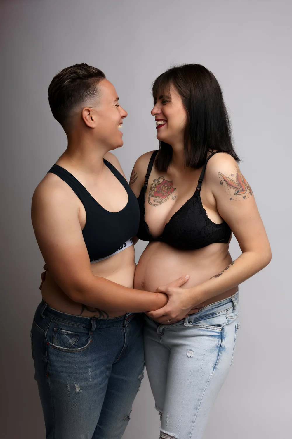 Couple en studio en jean et brassière noire, mains sur le ventre.
