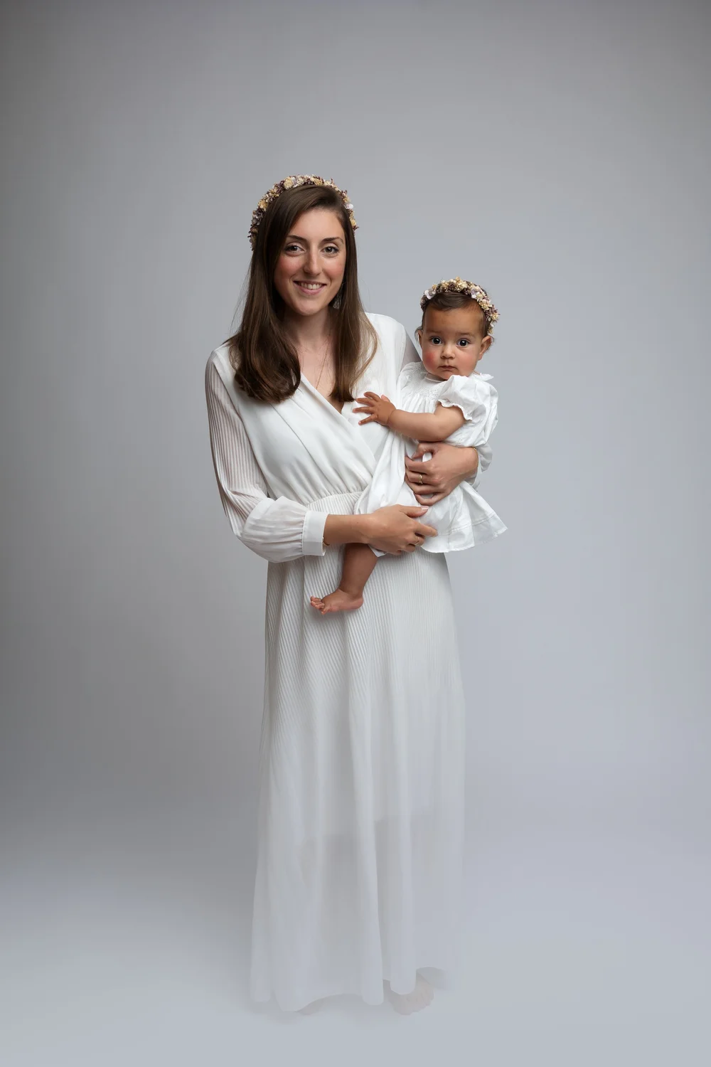Maman en robe blanche tenant son bébé, couronnes de fleurs, sur fond clair.