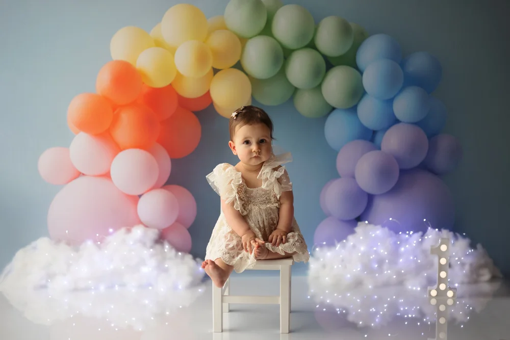 Bébé assise devant une arche de ballons arc-en-ciel et des nuages.