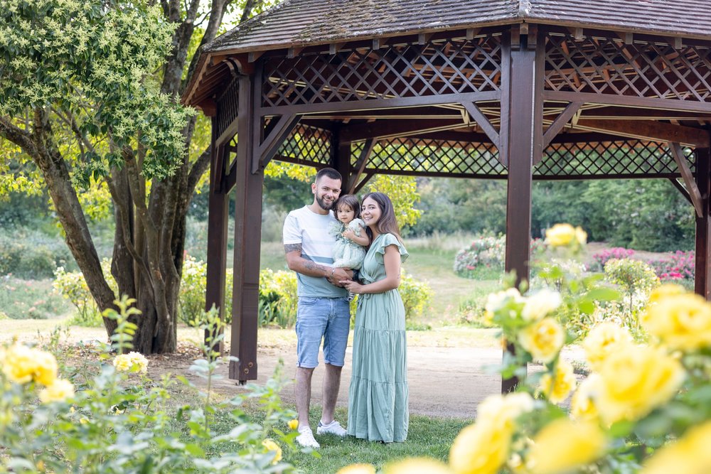 Couple et leur bébé devant un kiosque en bois, entouré de roses jaunes.