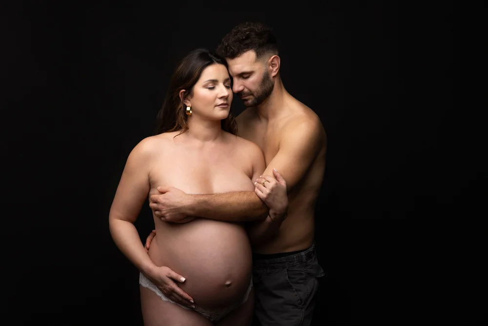 Couple en studio sur fond noir, mains sur le ventre.