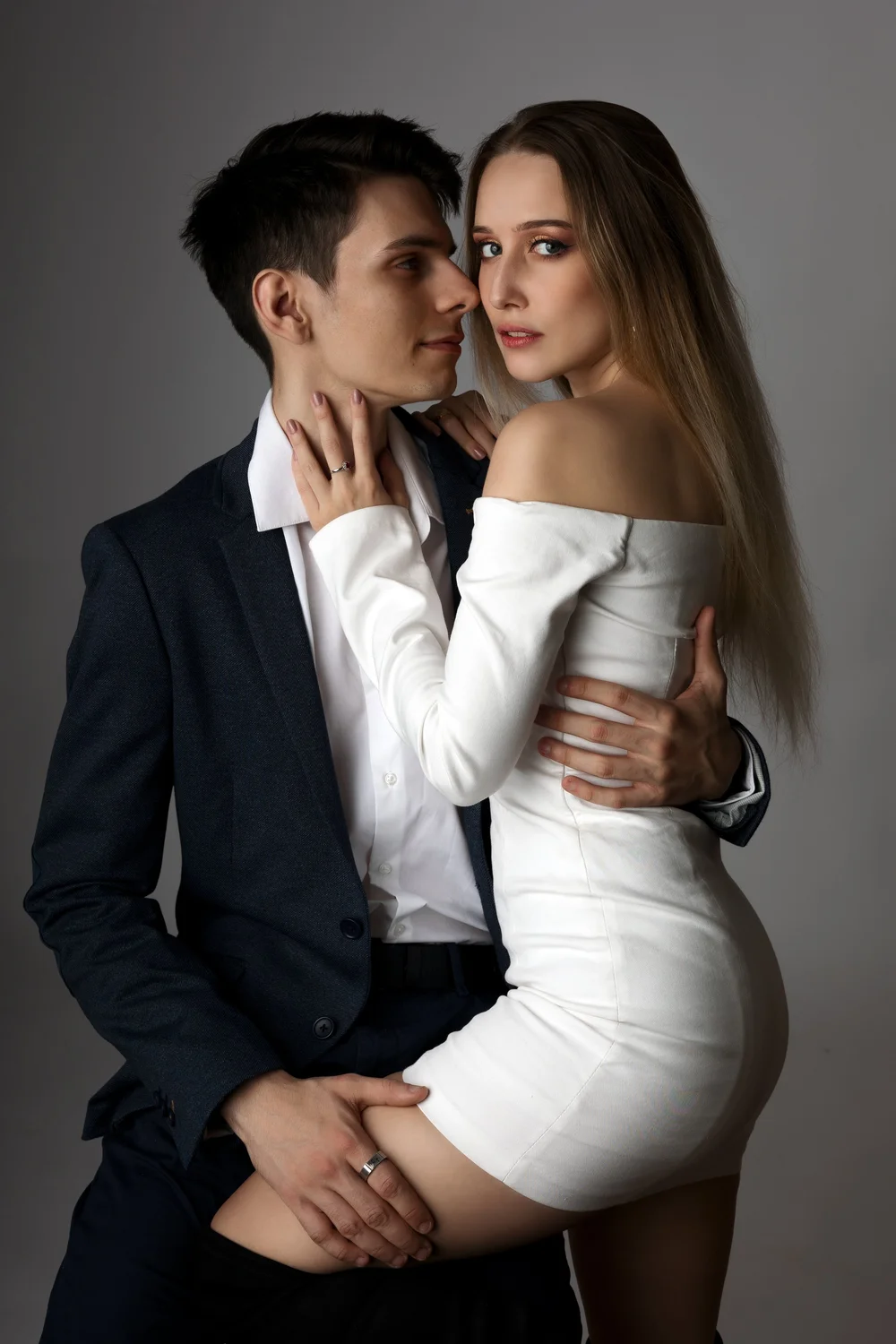 Couple enlacé en studio, lui en costume, elle en robe blanche.