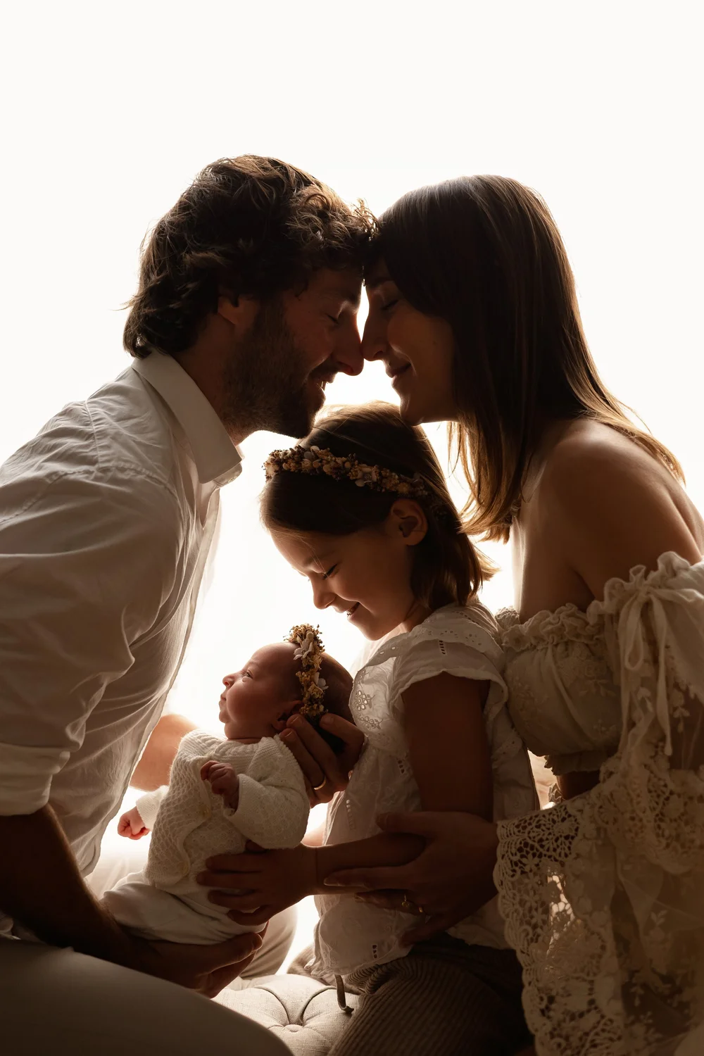 Parents front contre front au-dessus de leur fille et de leur nouveau-né, en contre-jour.