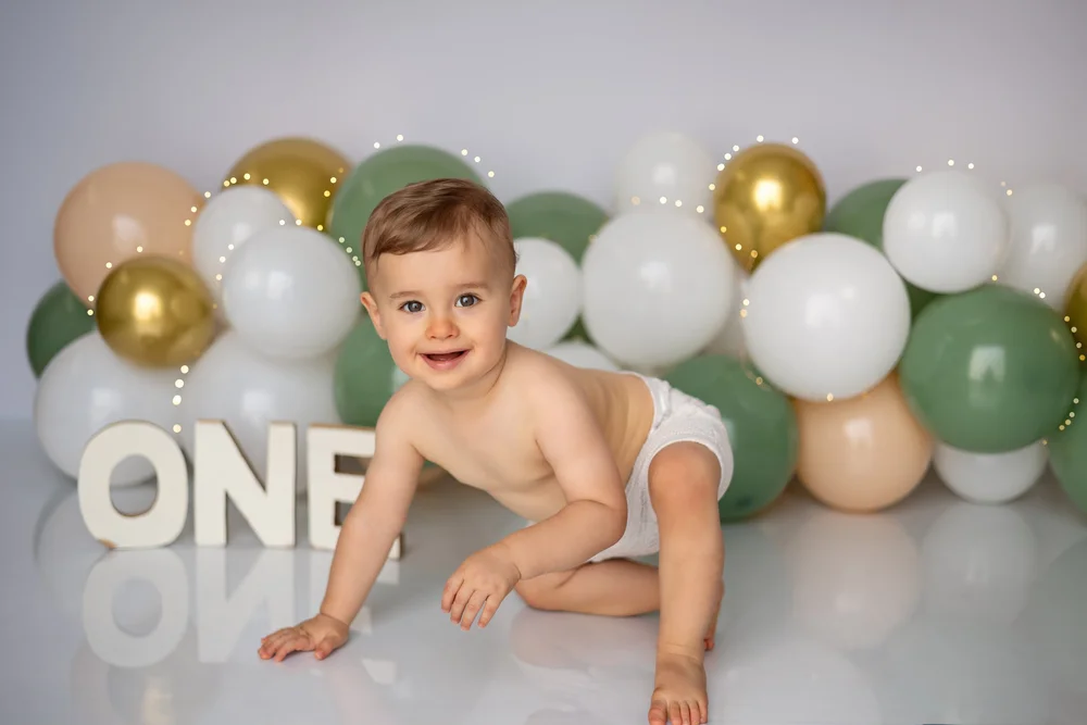 Bébé à quatre pattes devant une arche de ballons verts et dorés, lettres « ONE ».