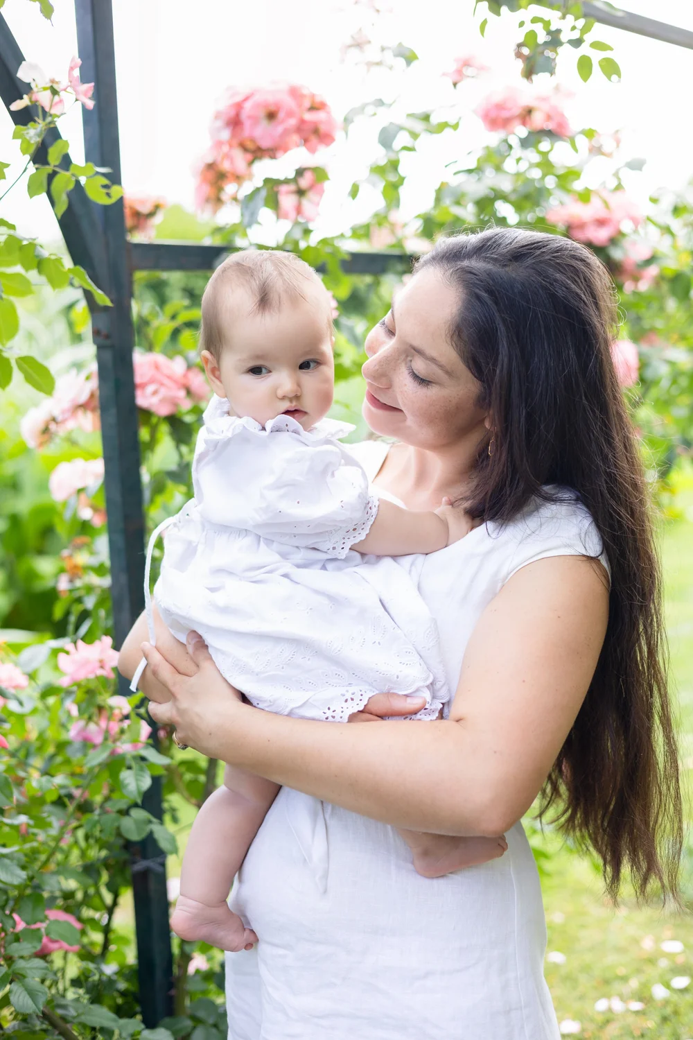 Maman en robe blanche portant son bébé sous une arche de roses.