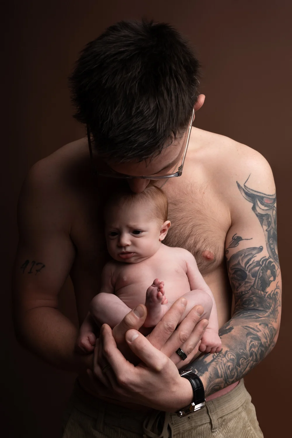 Papa torse nu tenant son nouveau-né, sur fond brun.