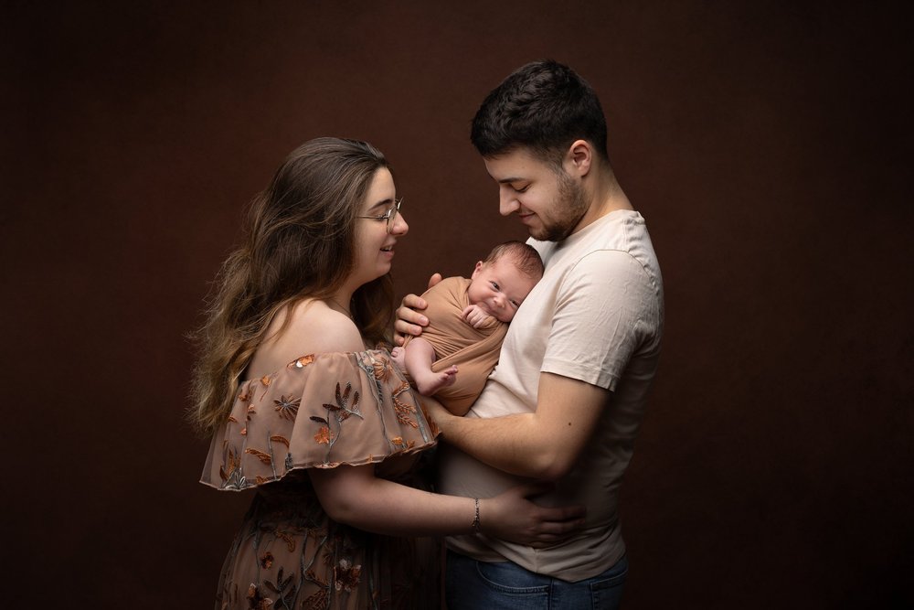 Parents en studio sur fond brun, le nouveau-né dans les bras.