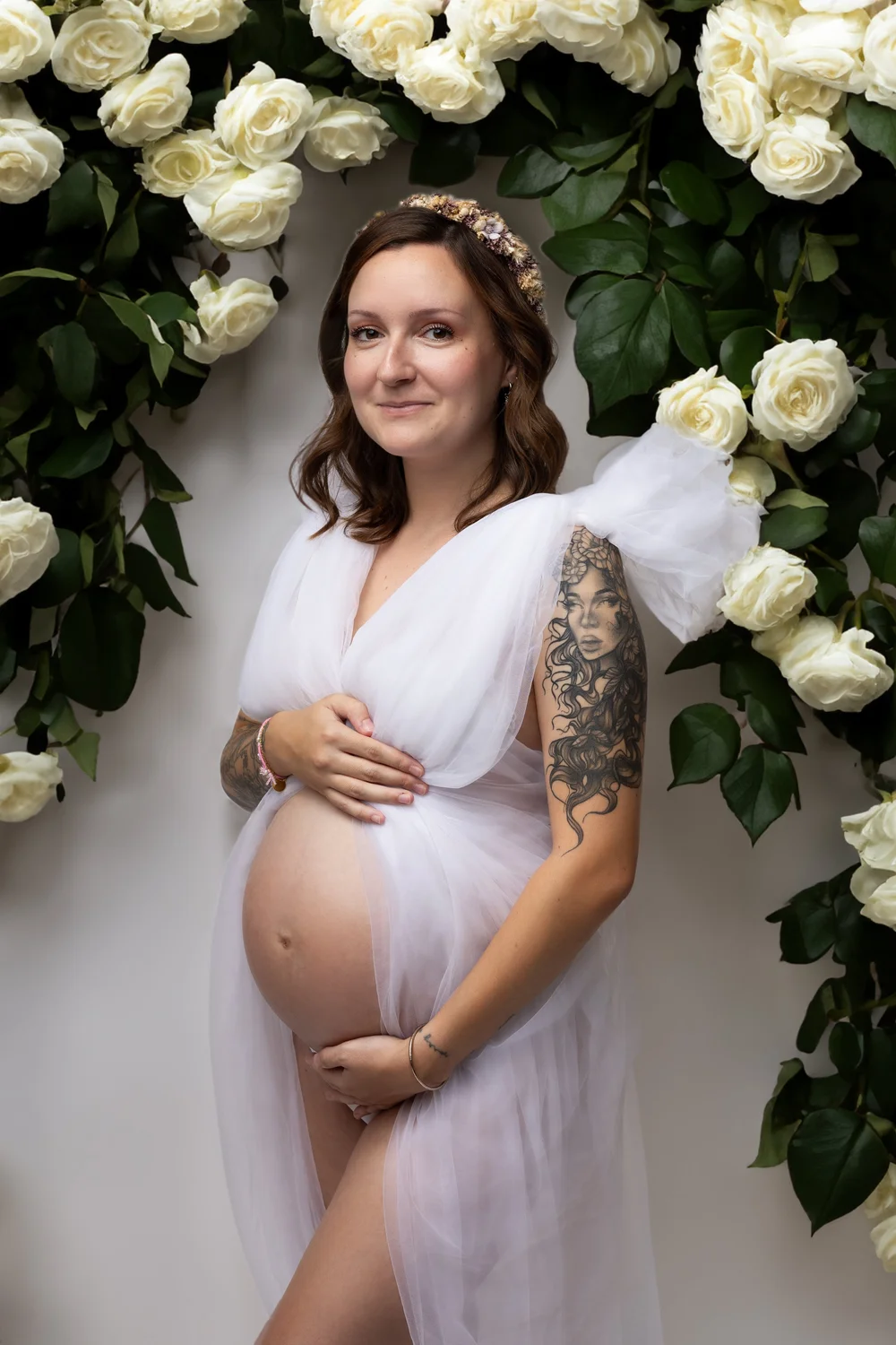 Femme enceinte en robe blanche transparente devant un massif de roses blanches.