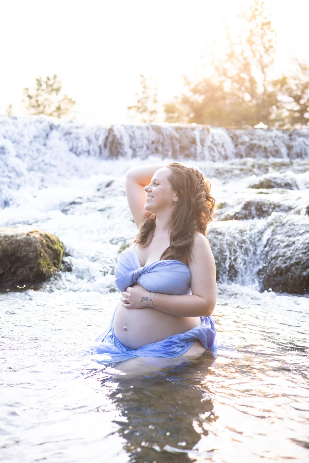 Femme enceinte les pieds dans l’eau devant une petite cascade, drapée de bleu.