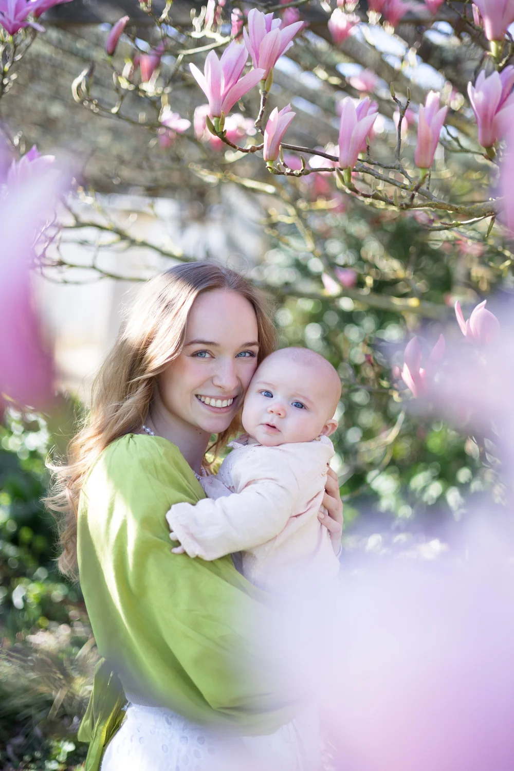 Maman en pull vert tenant son bébé sous un magnolia en fleurs.