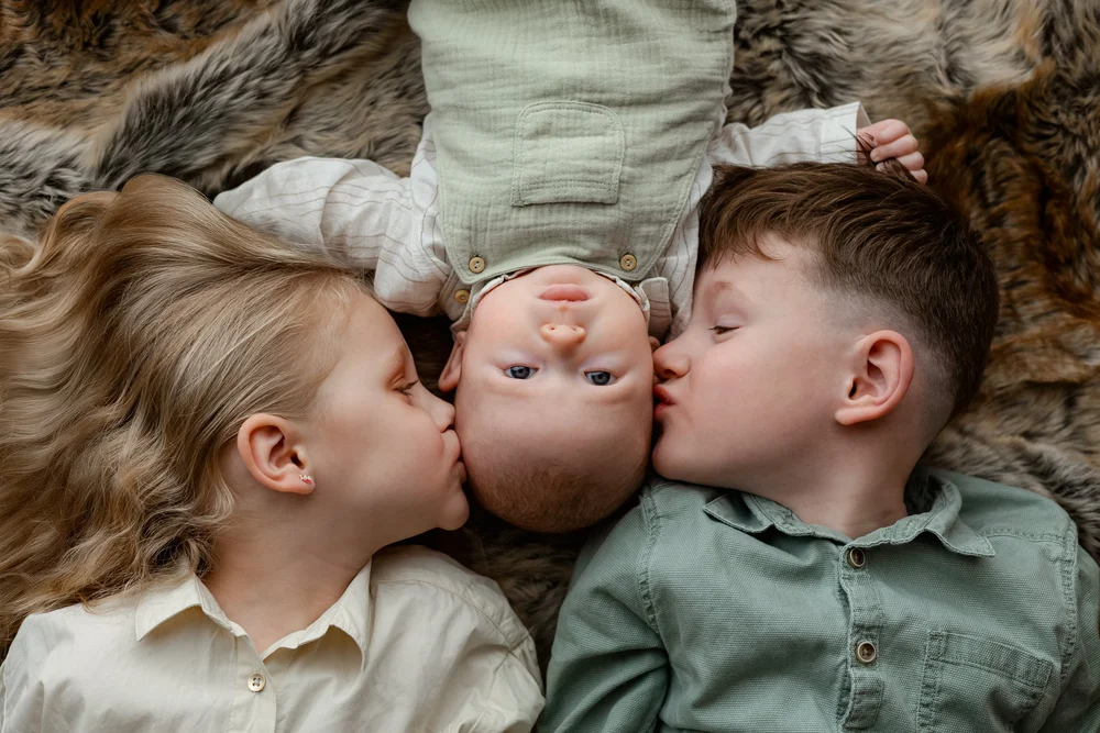 Deux enfants embrassant leur petit frère, allongés tête-bêche sur une fourrure.