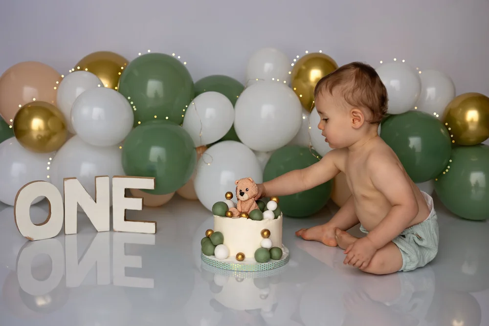 Bébé touchant son gâteau devant des ballons verts, lettres « ONE ».