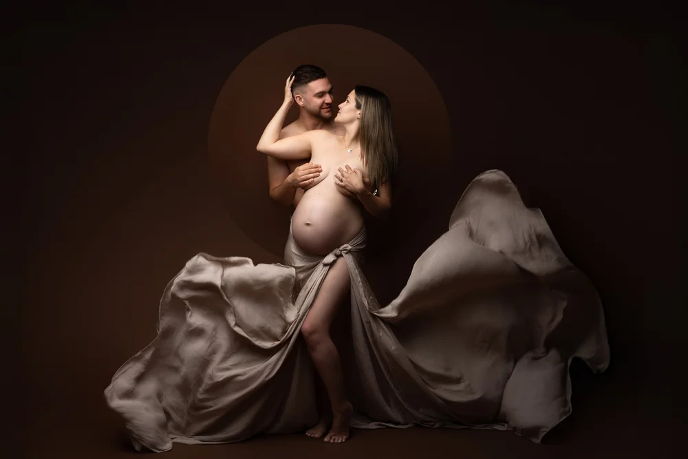 Femme enceinte drapée d’un voile beige avec son conjoint, en studio sur fond brun.